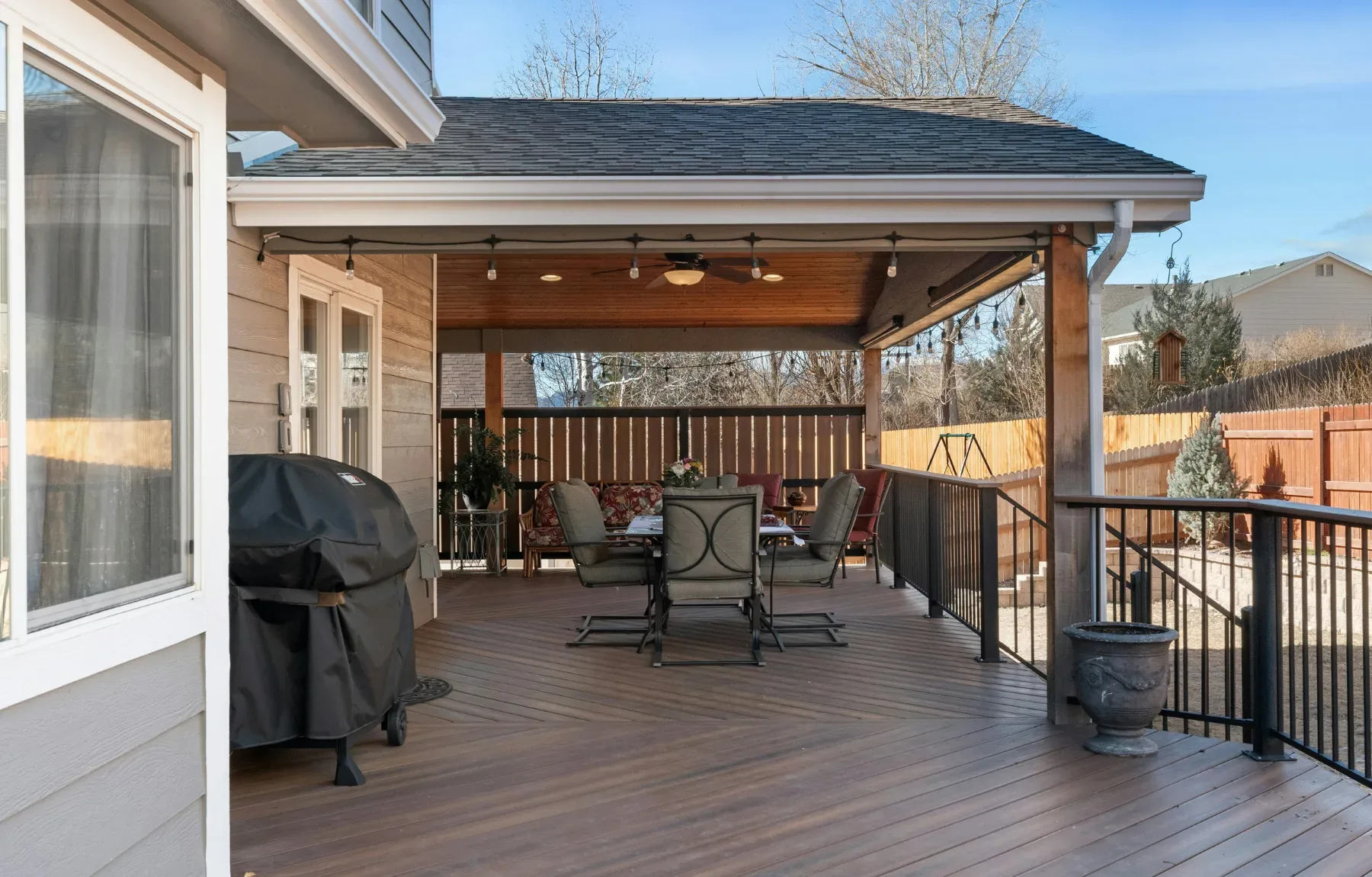 A covered composite deck attached to a house, with a shingled roof, wood-paneled ceiling, string lights, and a ceiling fan ov