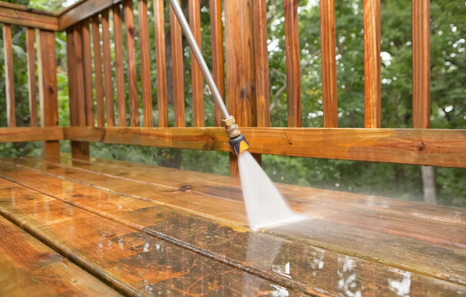 A pressure washer wand sprays a fan-shaped jet of water across a reddish-brown wooden deck, with wet boards glistening in the