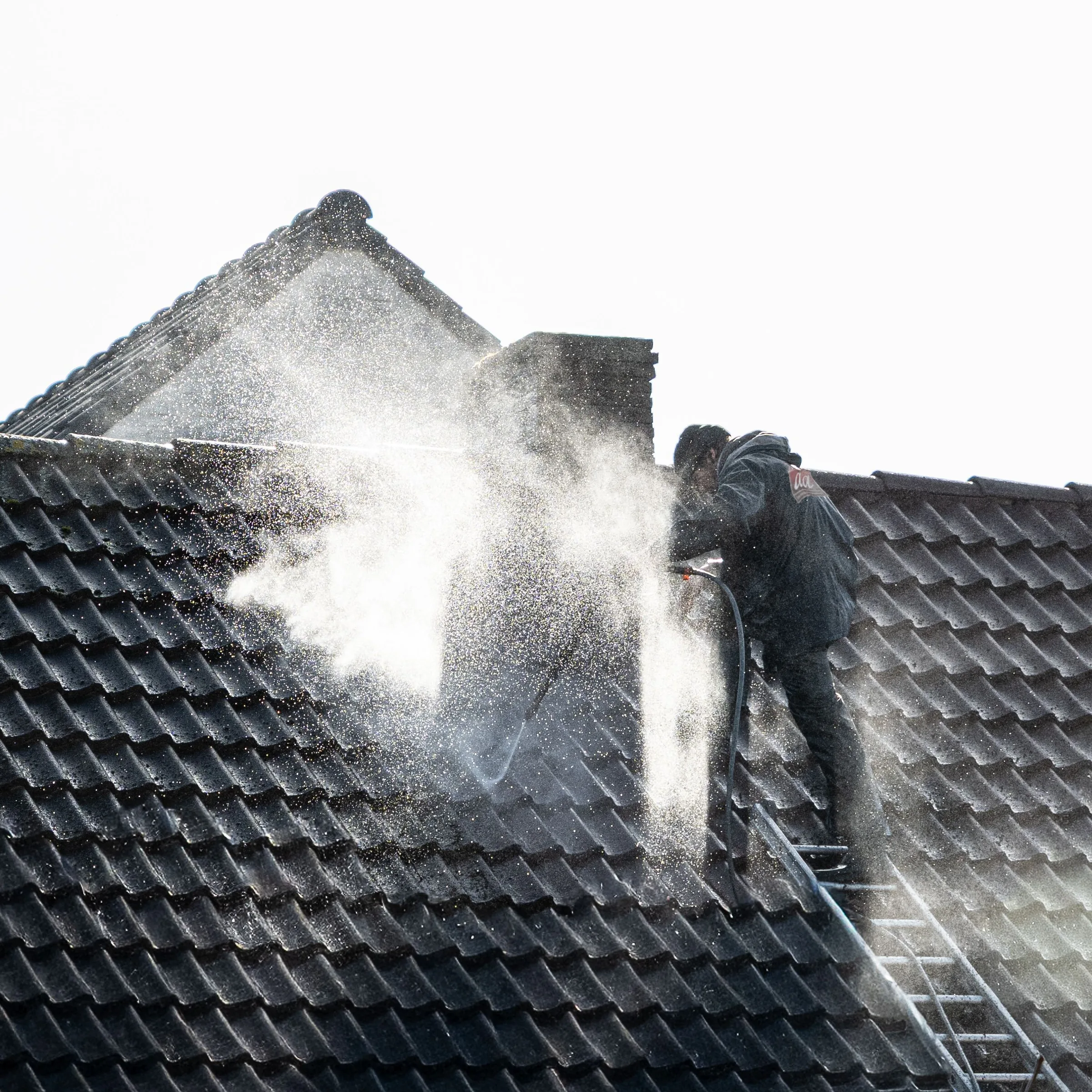 Worker pressure washing a dark tile roof with high-pressure water spray.