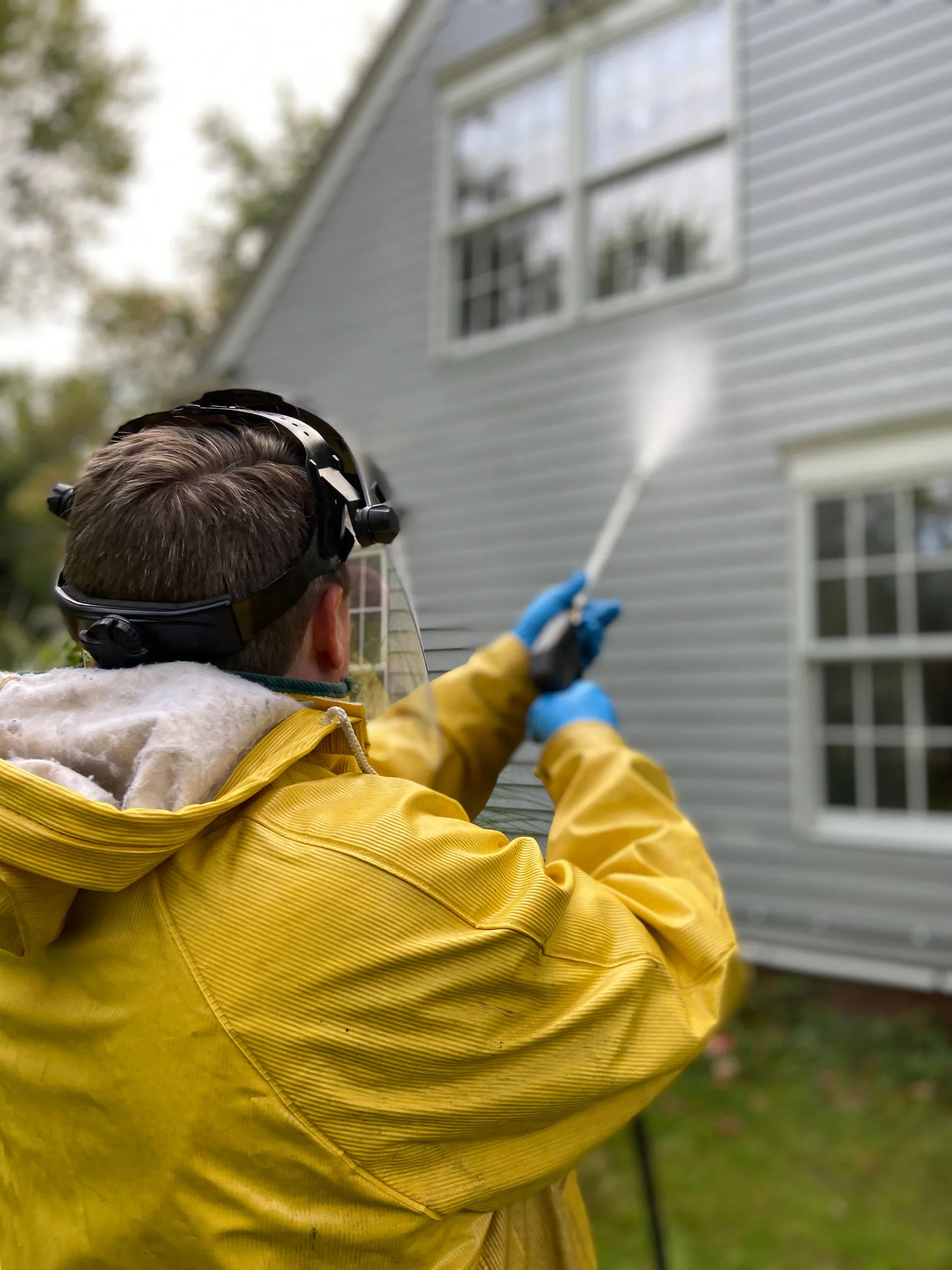 Worker in yellow jacket and protective gear pressure washing a gray house exterior.